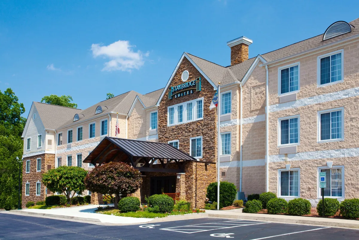 The image shows the hotel's exterior entrance, featuring an inviting entrance awning, a modern stone facade, and well-maintained landscaped greenery. The prominent hotel signage welcomes guests, and the front area exudes a professional yet warm atmosphere suitable for arrivals and business travelers.