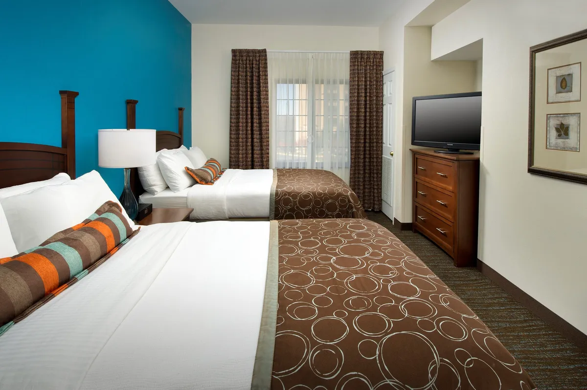 The image displays a welcoming second bedroom featuring two beds with decorative pillows, a colorful accent wall, and wooden furniture. The setting is comfortable and inviting—perfect for families or friends sharing a stay. The room layout suggests space and togetherness while maintaining a warm, restful vibe.