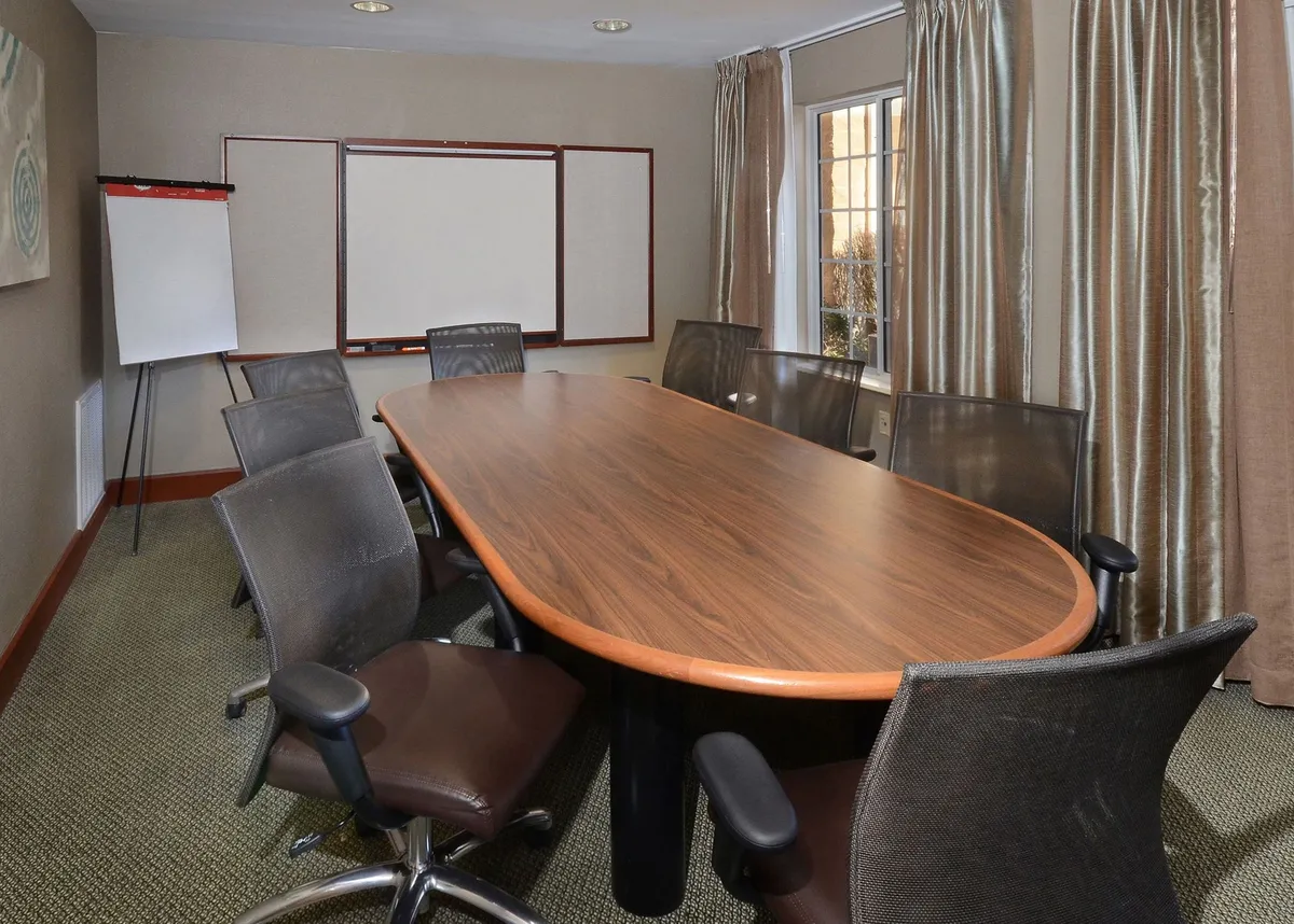 A well-lit business center featuring a long oval conference table surrounded by comfortable office chairs, a whiteboard for presentations, and large curtained windows letting in natural light. The space is set up for productivity, with a layout suitable for group meetings or quiet work.