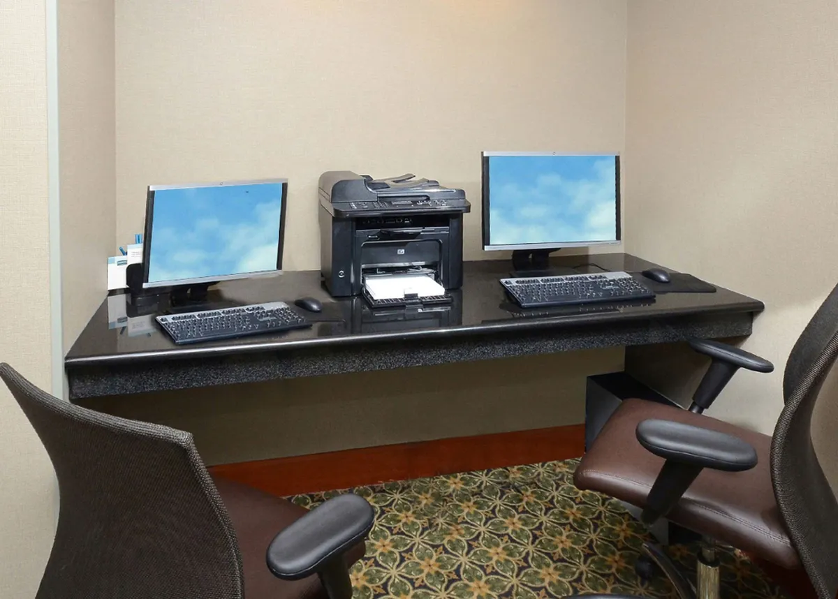 A dedicated workspace is depicted, featuring two computer monitors, a printer, and a spacious desk surface. The professional and tidy arrangement suggests a productive environment, ideal for catching up on work, planning meetings, or handling business tasks while traveling. The lighting and setup reinforce a sense of efficiency and focus.