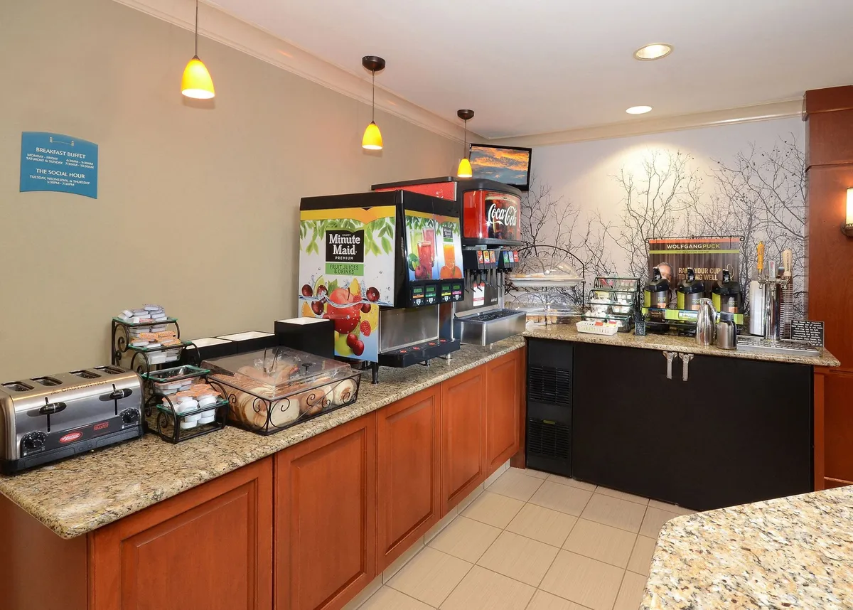 A bright breakfast bar featuring beverage dispensers, a tempting pastry display, and granite counters set in a communal dining space. The mood is casual and social, inviting guests to enjoy a complimentary breakfast or snack.