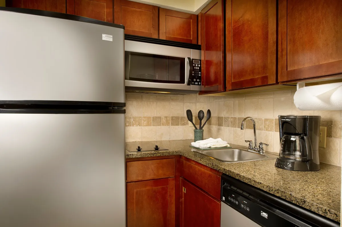 A closer look at the kitchen’s main utilities, including a stainless steel refrigerator, microwave, sink with modern faucet, and a coffee maker on a granite counter. The scene has a functional yet cozy vibe, suggesting both convenience and comfort.