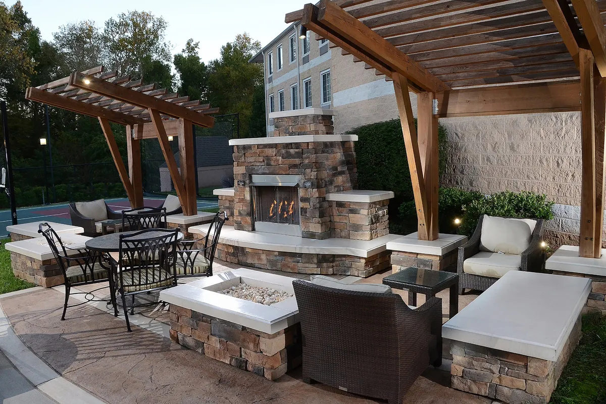 The image highlights a pergola-covered patio with a cozy fireplace, comfortable seating, and a table set for outdoor dining or casual gatherings. String lights and lush surroundings create a warm, communal atmosphere suited for evening relaxation or celebrations.