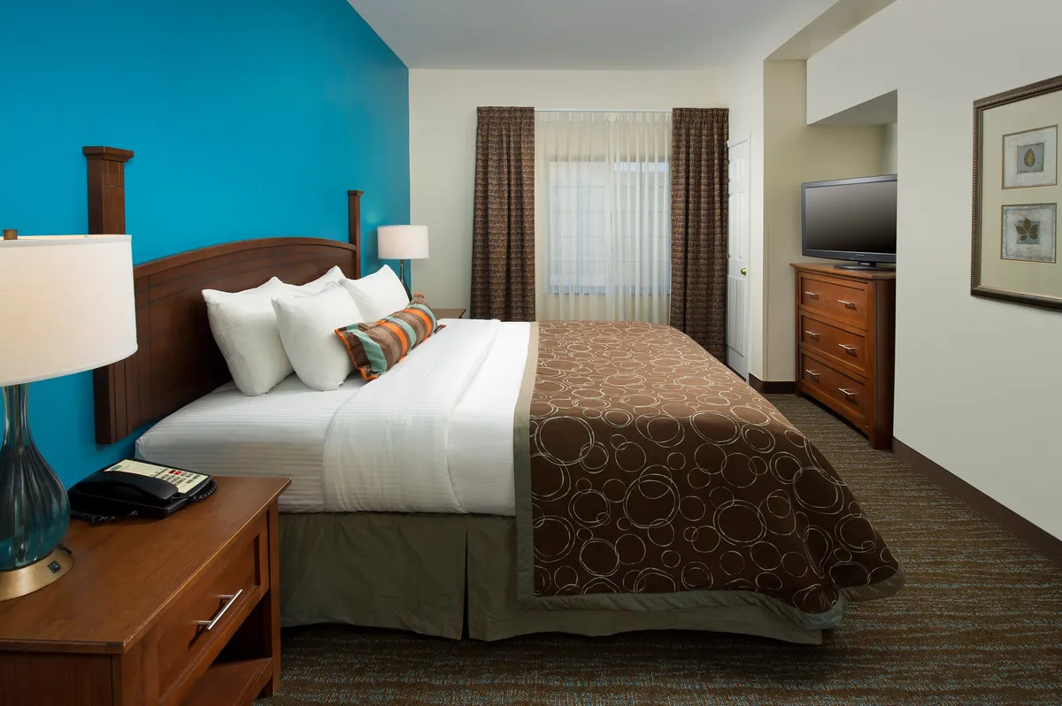 This image features a bedroom with a comfortable bed, a blue accent wall, bedside lamp, drapes, and a television. The room exudes a sense of coziness and relaxation, with sunlight streaming through the curtains, showcasing its tranquil décor and inviting atmosphere.